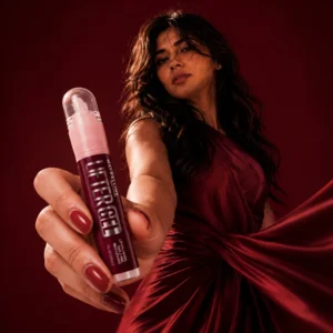 Woman in a burgundy satin dress holding Maybelline Lifter Gel Lip Gloss in a luxury beauty advertisement with dramatic burgundy background and professional studio lighting.